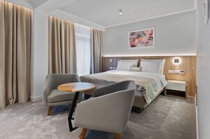 A modern hotel room with a large bed, two armchairs, a small round table, and a nightstand with a lamp.