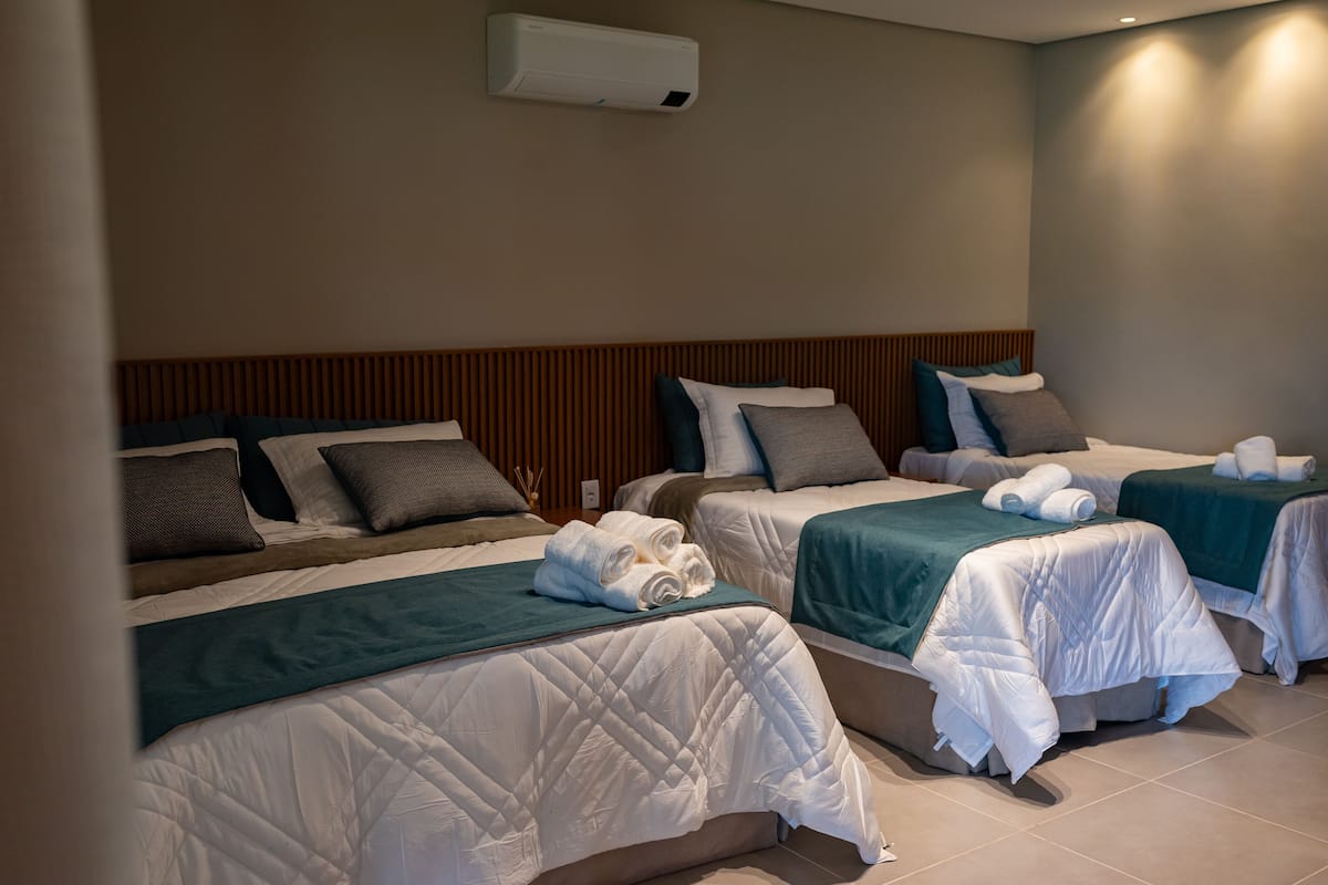 A hotel room with three beds, each with a quilted white duvet, teal bedspreads, and neatly folded towels.