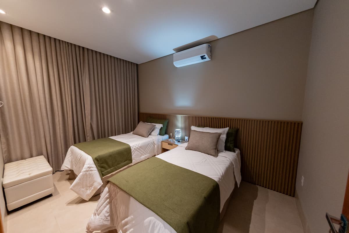 A hotel room with two beds, a bedside table, an air conditioning unit, and a window with blinds.