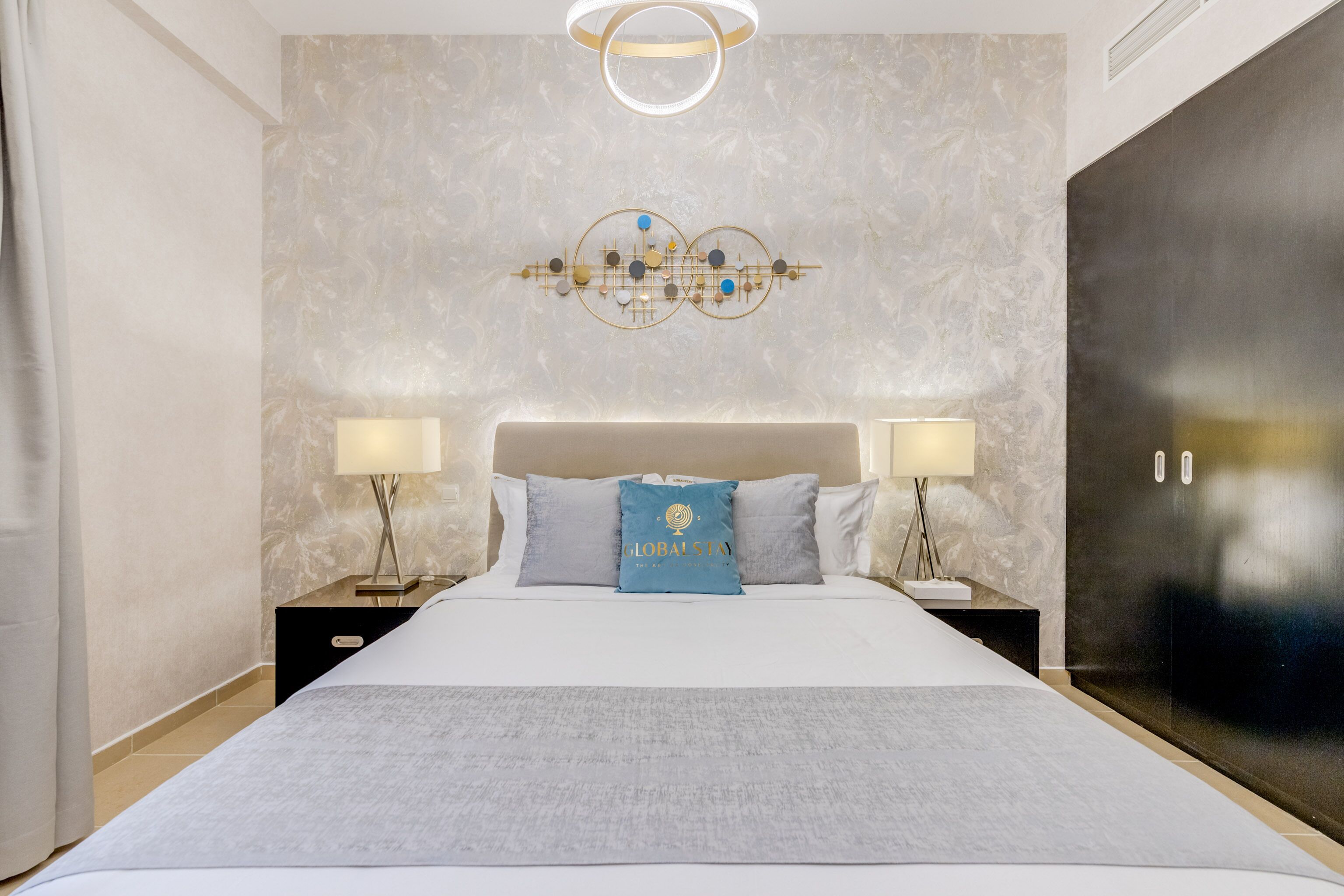 Une chambre d’hôtel moderne avec un grand lit, deux tables de chevet avec des lampes, un élément décoratif mural et une armoire sombre.