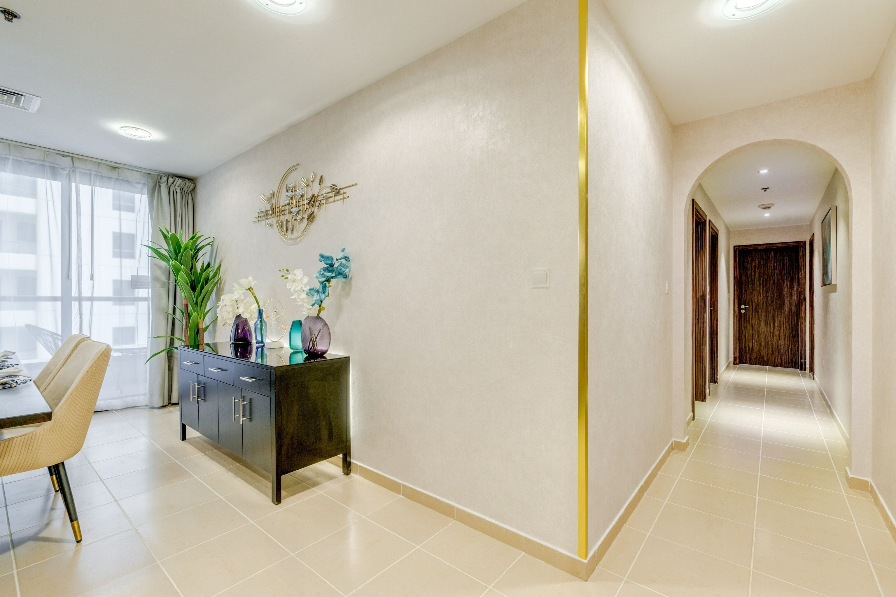 A modern interior with a dining area, a dark wooden cabinet with decorative items, a large plant, and a hallway with an arched doorway.
