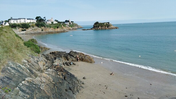A coastal landscape with a rocky beach, a small island, and buildings in the background.