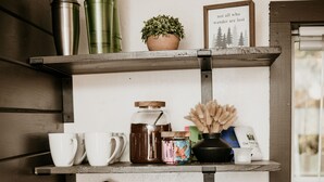 Una cucina con una macchina da caffè, uno scaffale con tazze, un barattolo e una pianta in vaso.
