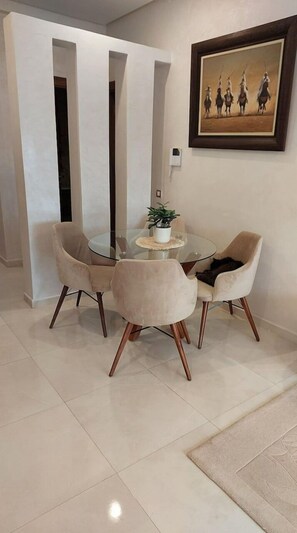 Uma área de jantar com mesa de vidro, rodeada por quatro cadeiras, um vaso de planta na mesa e um quadro na parede.