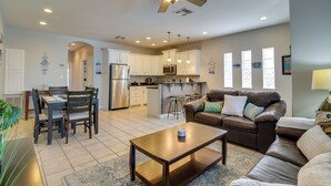A spacious living room with a brown leather sofa, a wooden coffee table, and a dining area with a wooden table and chairs. A kitchen with white cabinets and stainless steel appliances is visible in the background. - South Padre Island Condo w/ Shared Outdoor Pool! (South Padre Island)