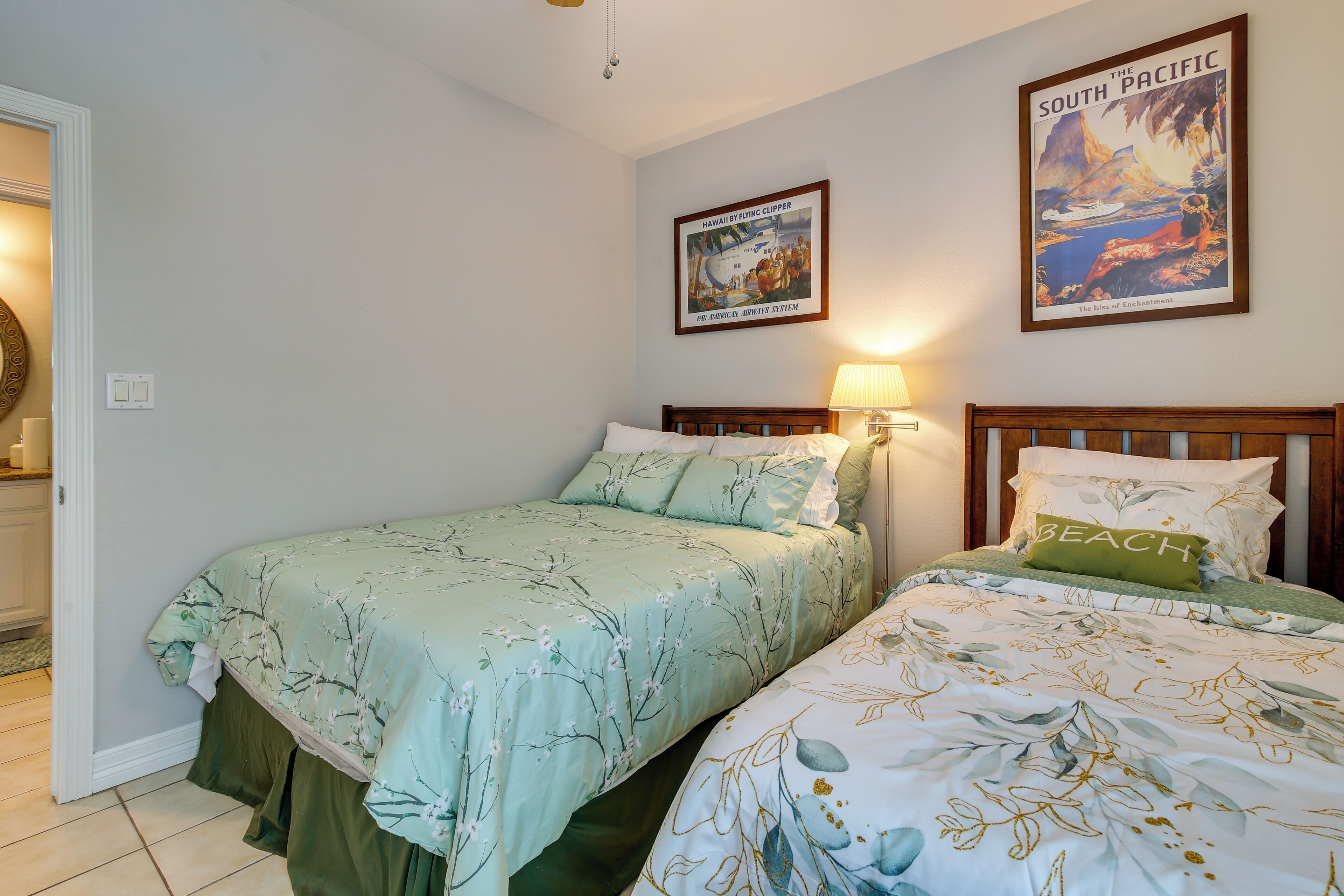 A bedroom with two beds, a framed picture of a tropical scene, and a lamp on the nightstand.