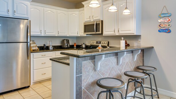A modern kitchen with white cabinets, stainless steel appliances, a granite countertop, and a bar area with stools. - South Padre Island Condo w/ Shared Outdoor Pool! (South Padre Island)