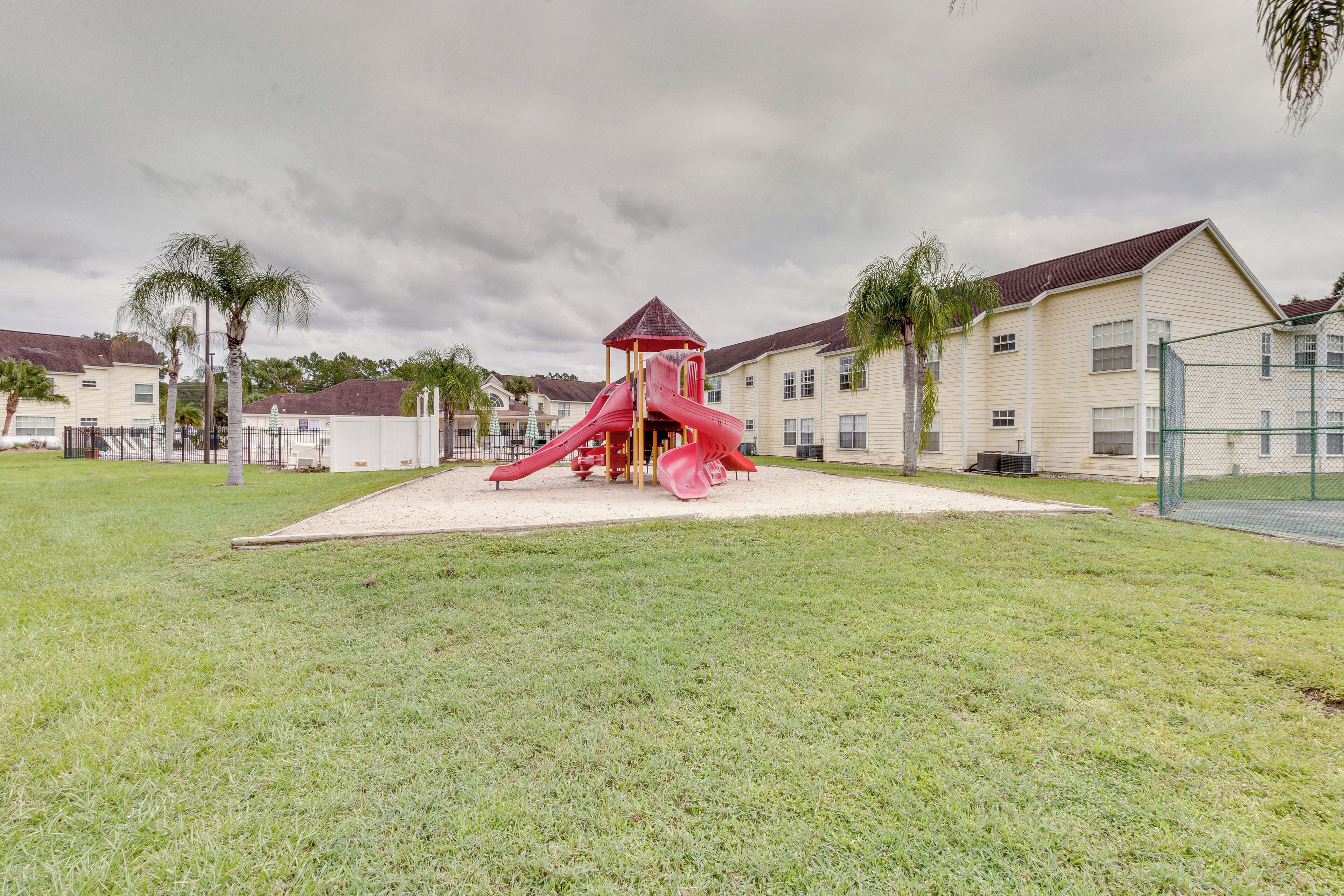 A playground with a red slide and a small shelter, surrounded by a grassy area and residential buildings in the background.