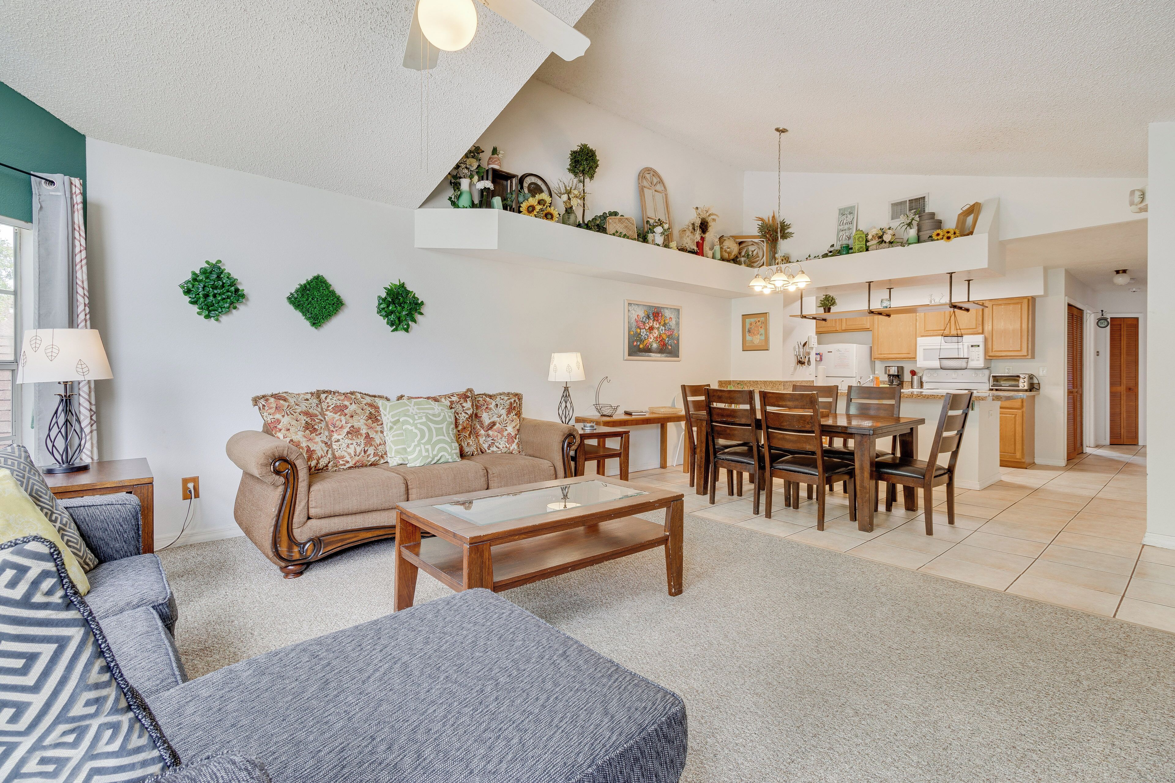 A spacious living area with a sofa, coffee table, and dining table set. A kitchen is visible in the background with wooden cabinets and a microwave.