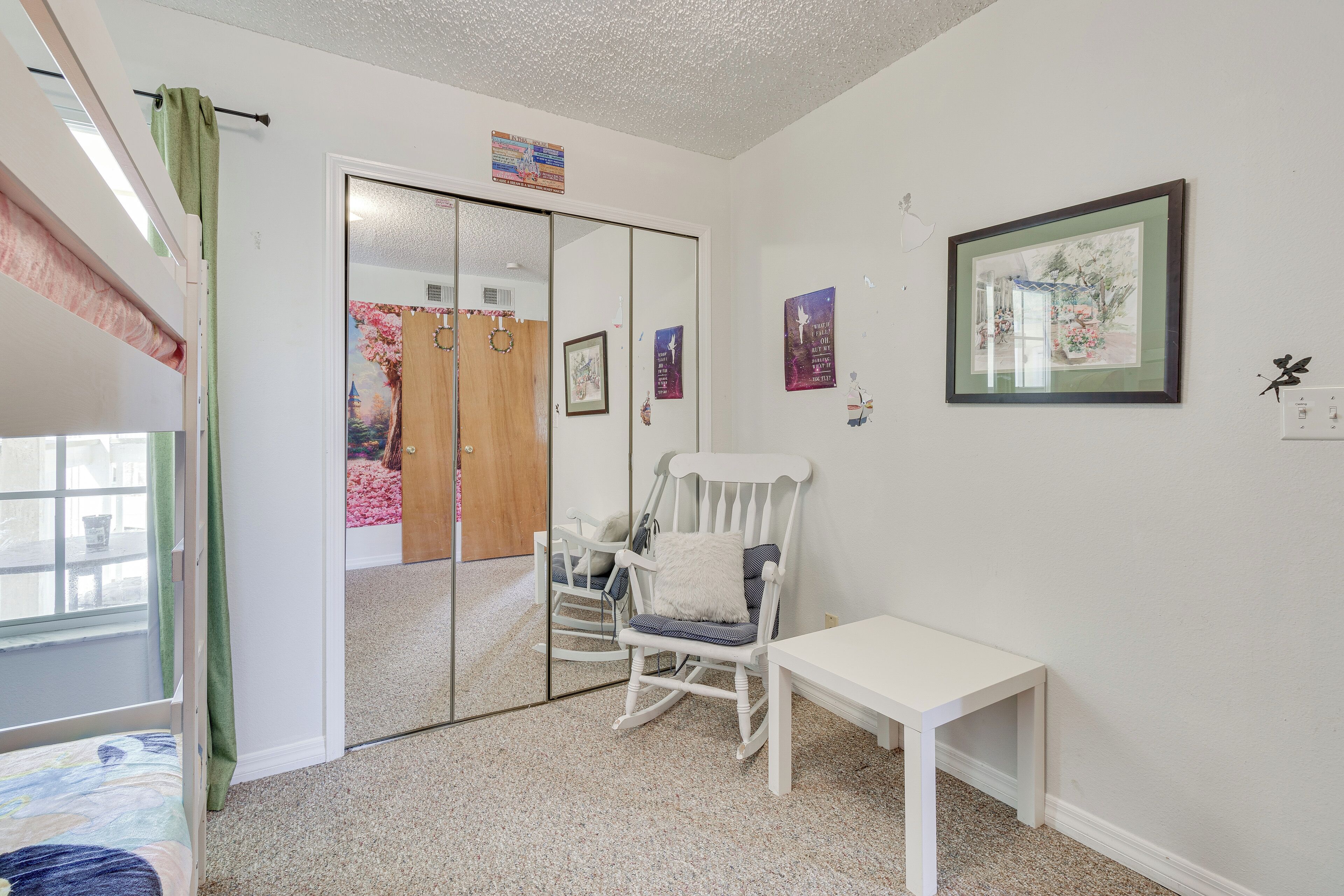 A bedroom with a bed, a rocking chair, a small table, and a mirror.
