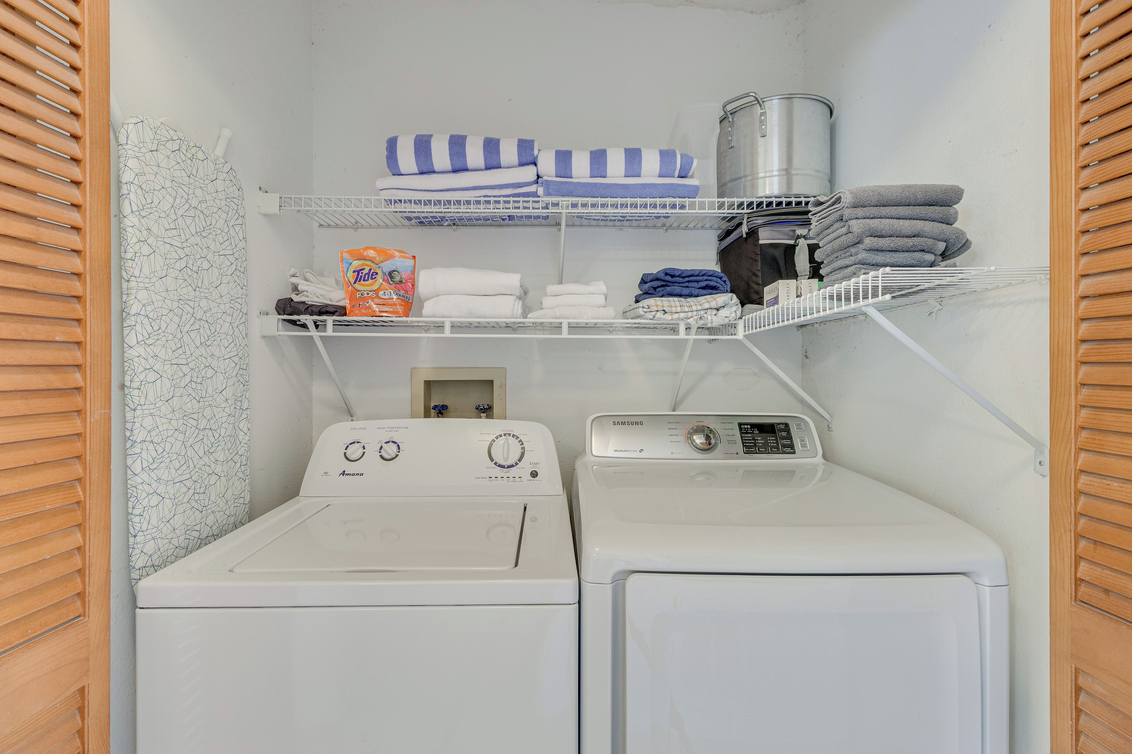 Un cuarto de lavandería con dos lavadoras, repisas con toallas dobladas y productos de limpieza, y un recipiente metálico.