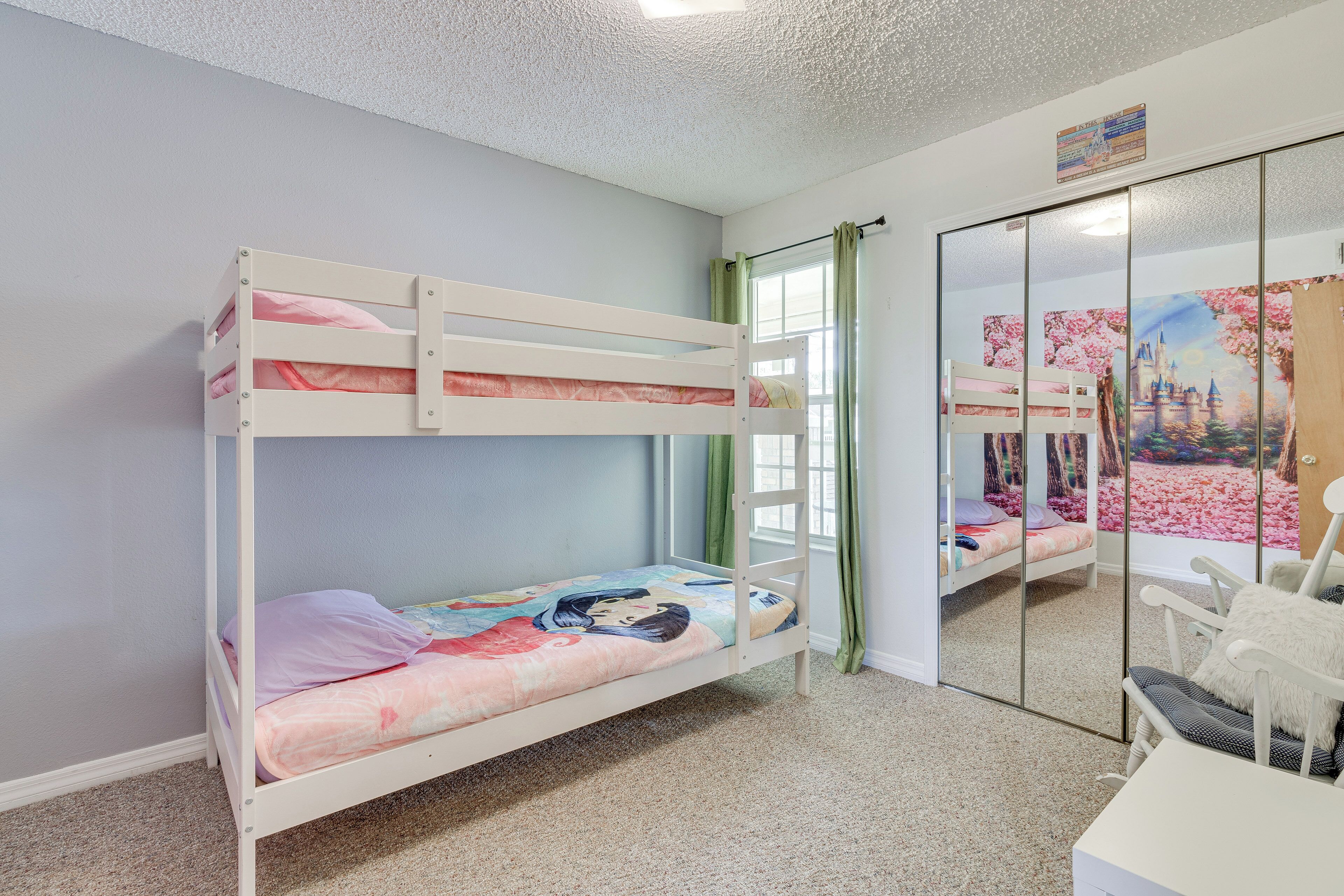 A bunk bed with a colorful bedspread, a white desk with a chair, and a large mirror reflecting a painting.