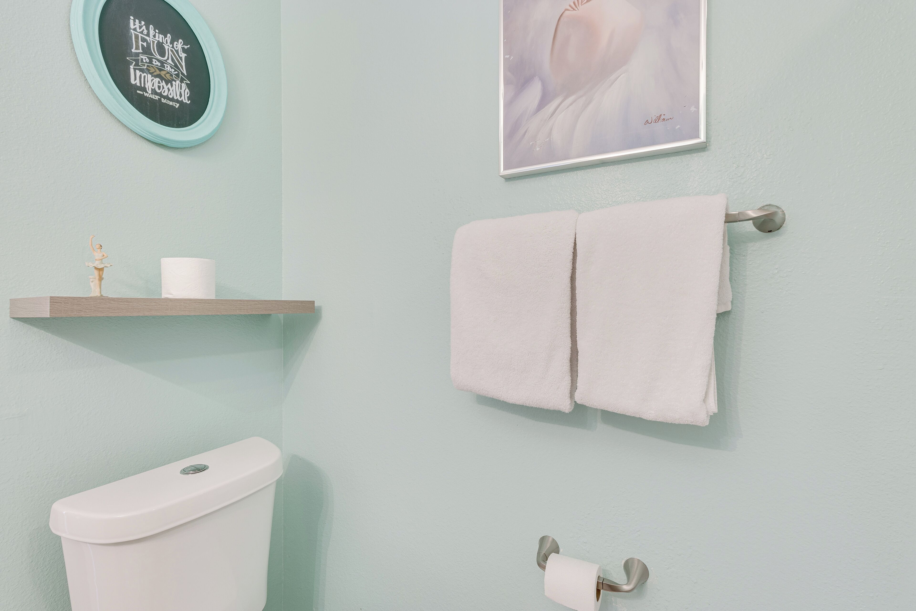 Un baño con inodoro, un perchero con dos toallas, un estante con un rollo de papel higiénico y un adorno, y una cita enmarcada en la pared.