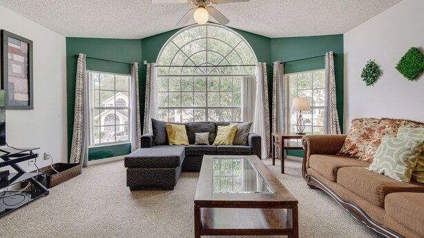 A living room with a sectional sofa, a glass-top coffee table, and a ceiling fan. - 6 Mi to Disney: Family-friendly Condo in Kissimmee (Kissimmee)