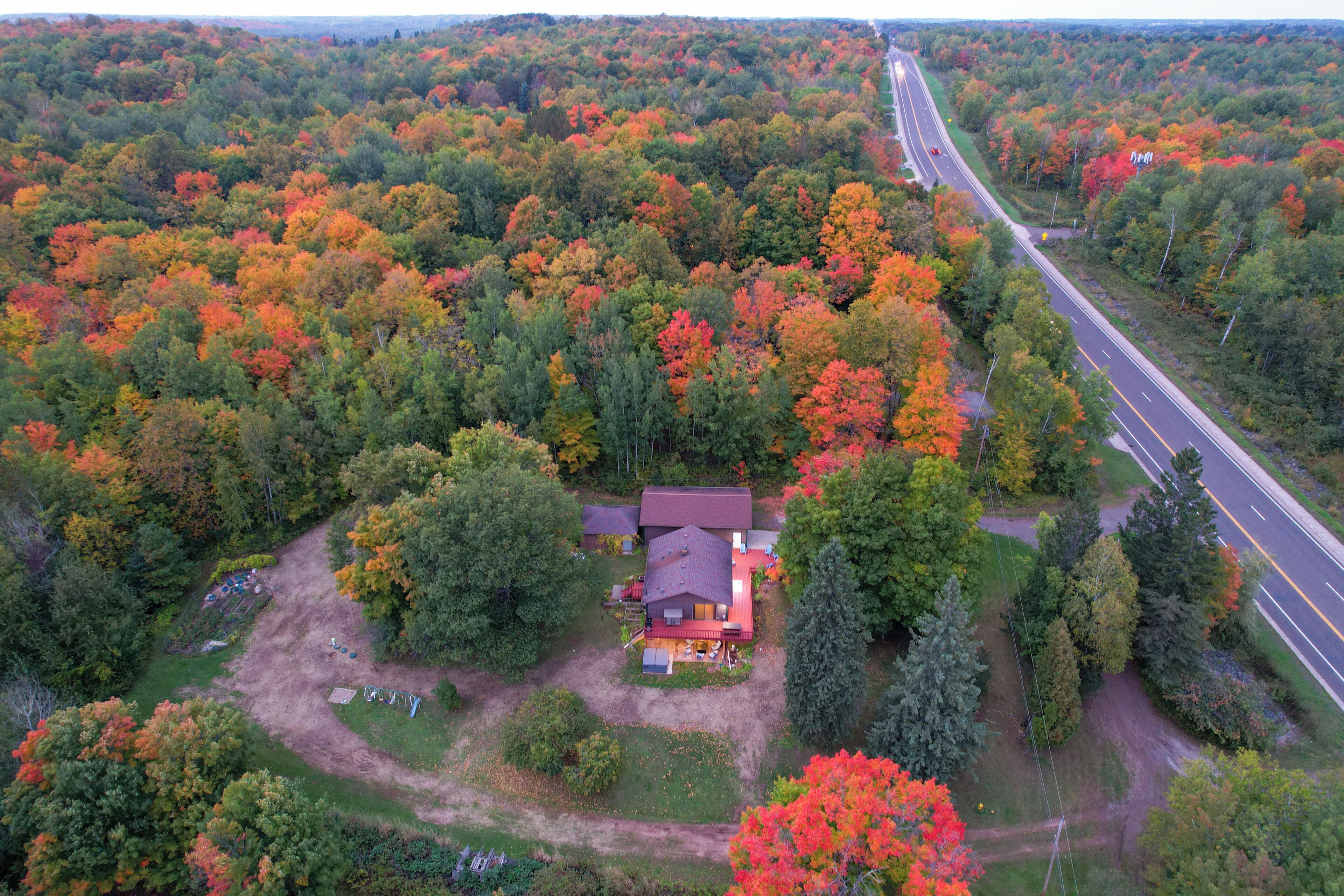 An aerial view of a house surrounded by a forest with autumn foliage. A road runs parallel to the property.
