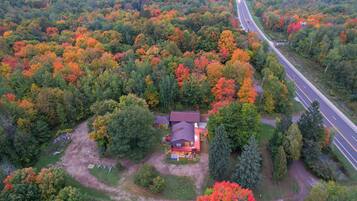 An aerial view of a house surrounded by a forest with autumn foliage. A road runs parallel to the property.