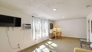 A room with a flat-screen TV mounted on the wall, a standing fan, a glass table, a sofa, and a window with curtains. - Old Orchard Beach Vacation Rental, Walk to Ocean! (Old Orchard Beach)