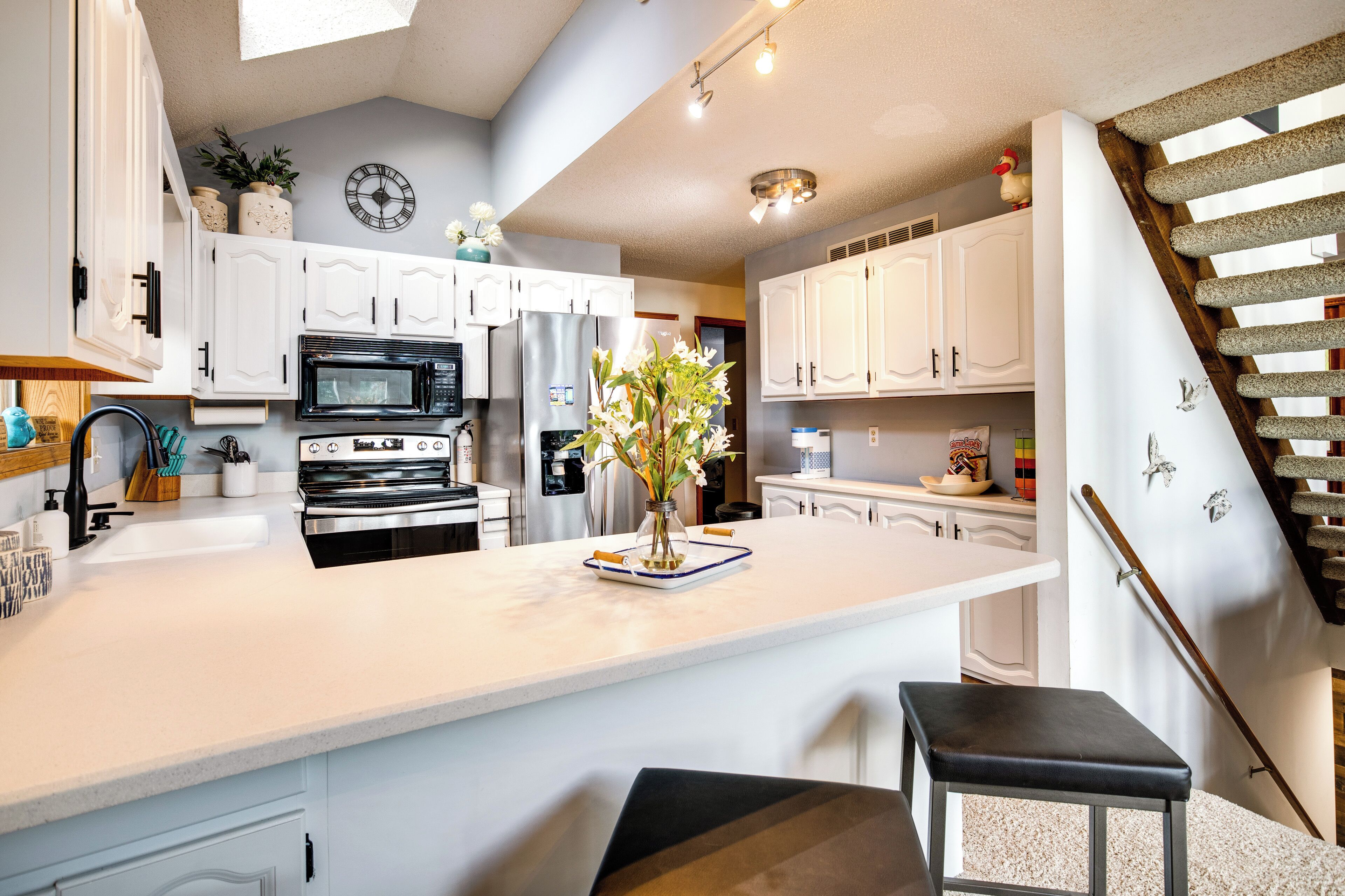 Una cocina moderna con armarios blancos, una isla central, un refrigerador de acero inoxidable y una escalera de madera.
