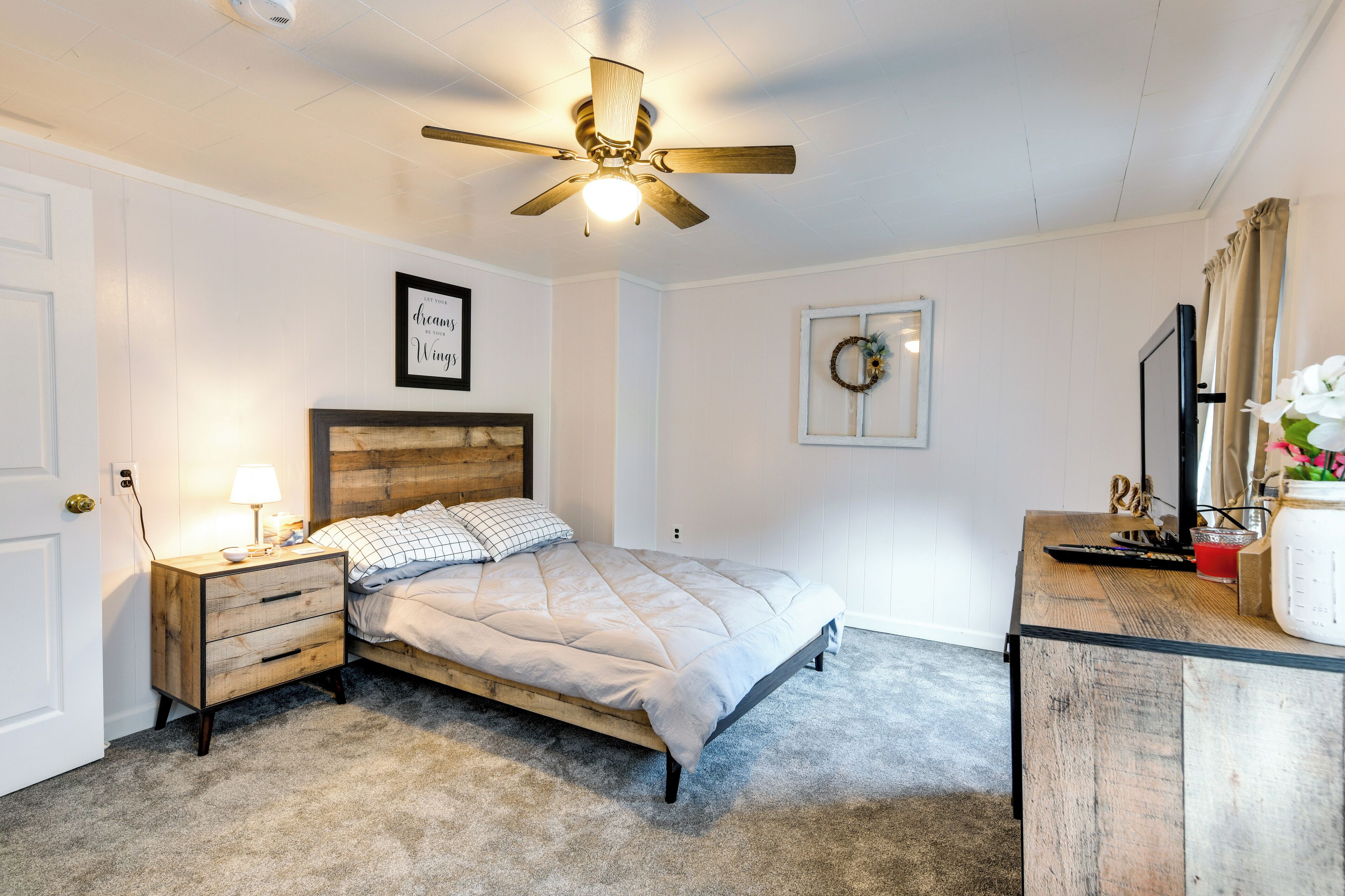 A bedroom with a bed, a nightstand, a desk, a ceiling fan, and a wall-mounted TV.