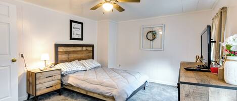A bedroom with a bed, a nightstand, a desk, a ceiling fan, and a wall-mounted TV.