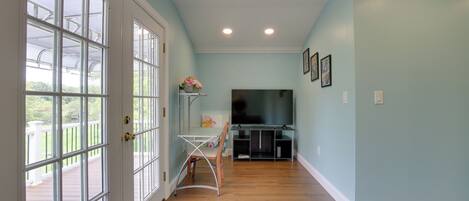 A hallway with a wooden floor, a white door with glass panels, a small table with a chair, and a TV stand with a flat-screen TV.