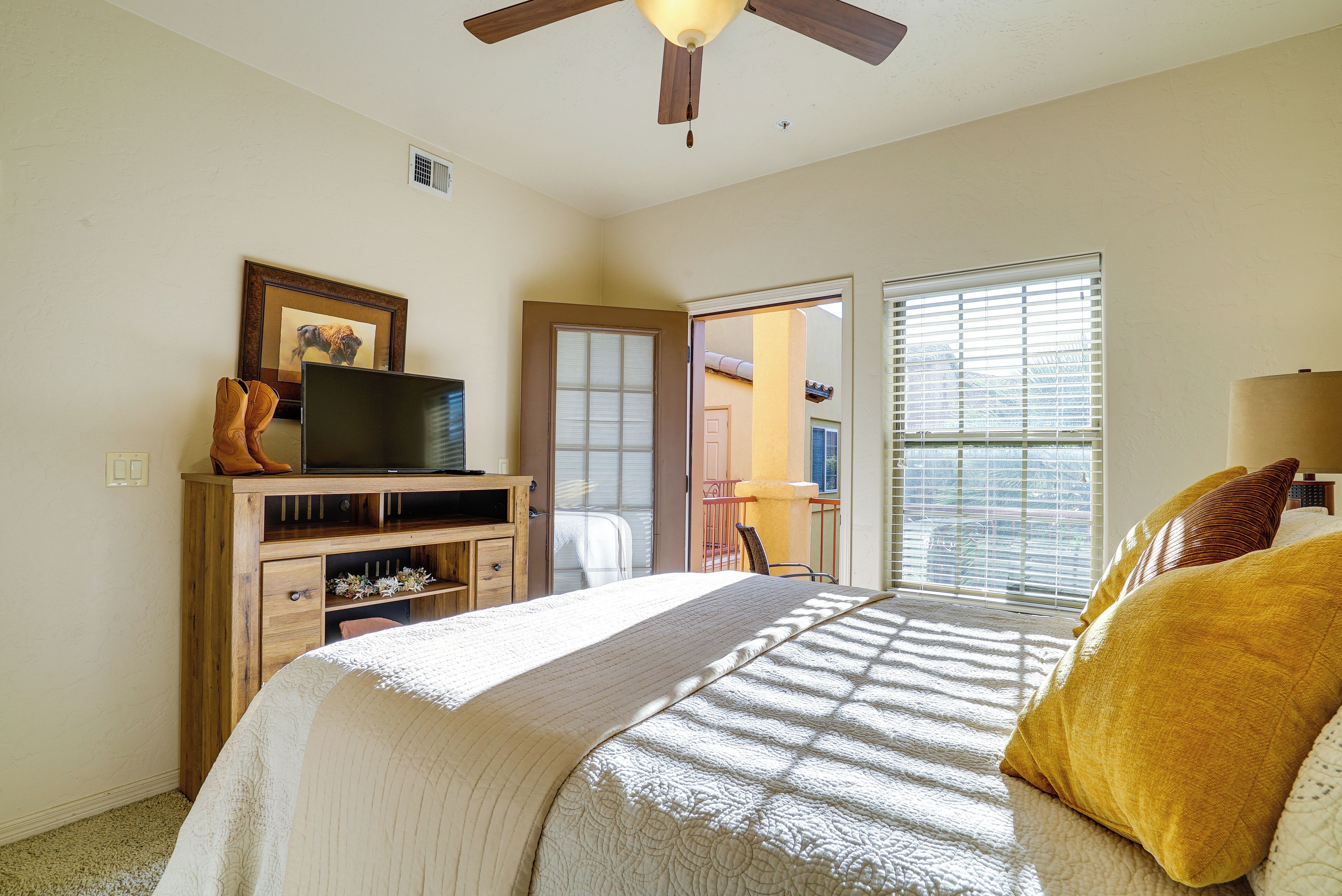 A bedroom with a bed, a television on a wooden stand, a door leading to another room, and a window with blinds.