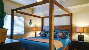 Een bed met hemelbed, met een houten frame, een blauwe quilt en meerdere kussens. Er staat een nachtkastje met een lamp, een klein krukje en een raam met jaloezieën.