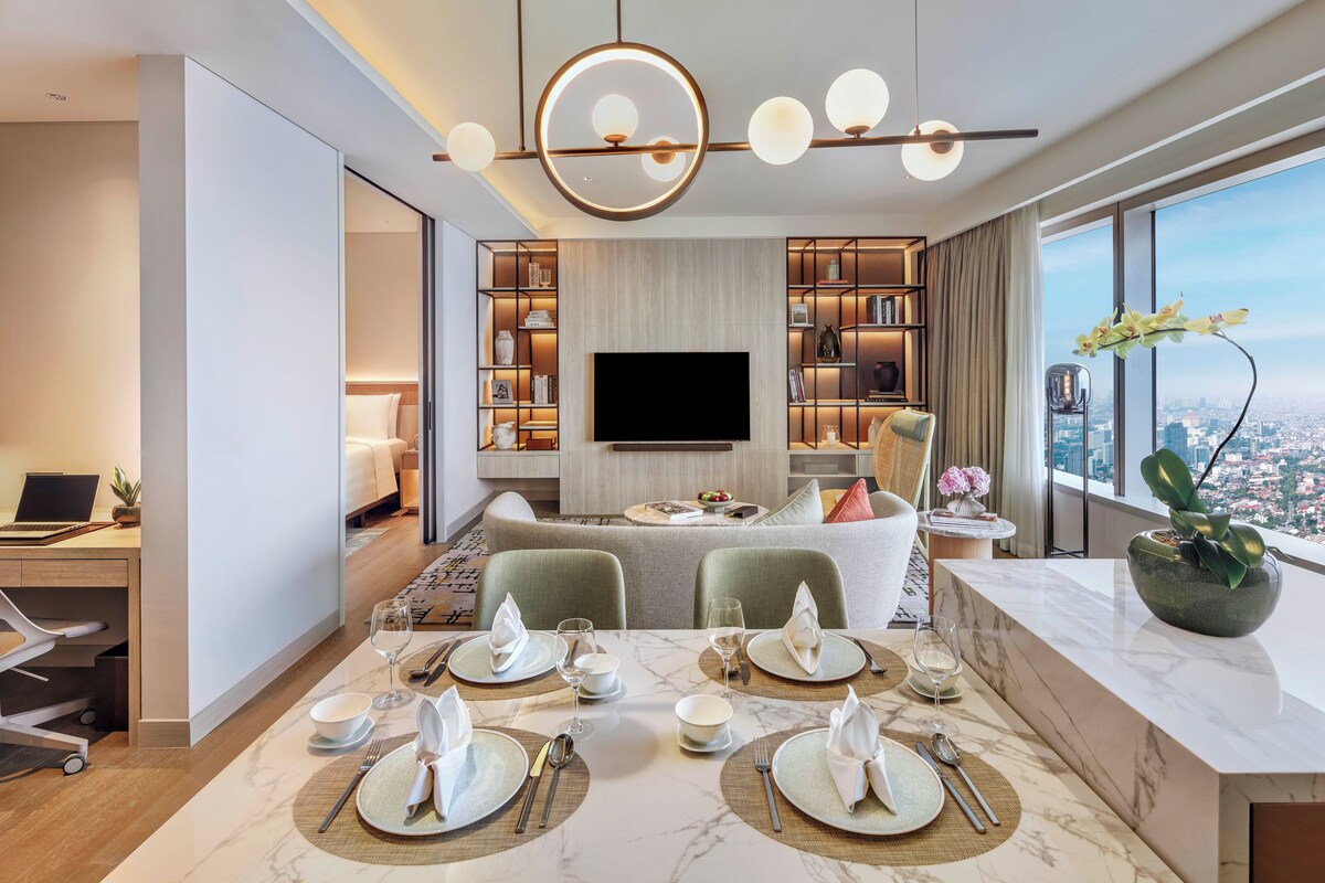 Habitación de hotel moderna con zona de comedor, televisor de pantalla plana, vista a la ciudad y un amplio ventanal.