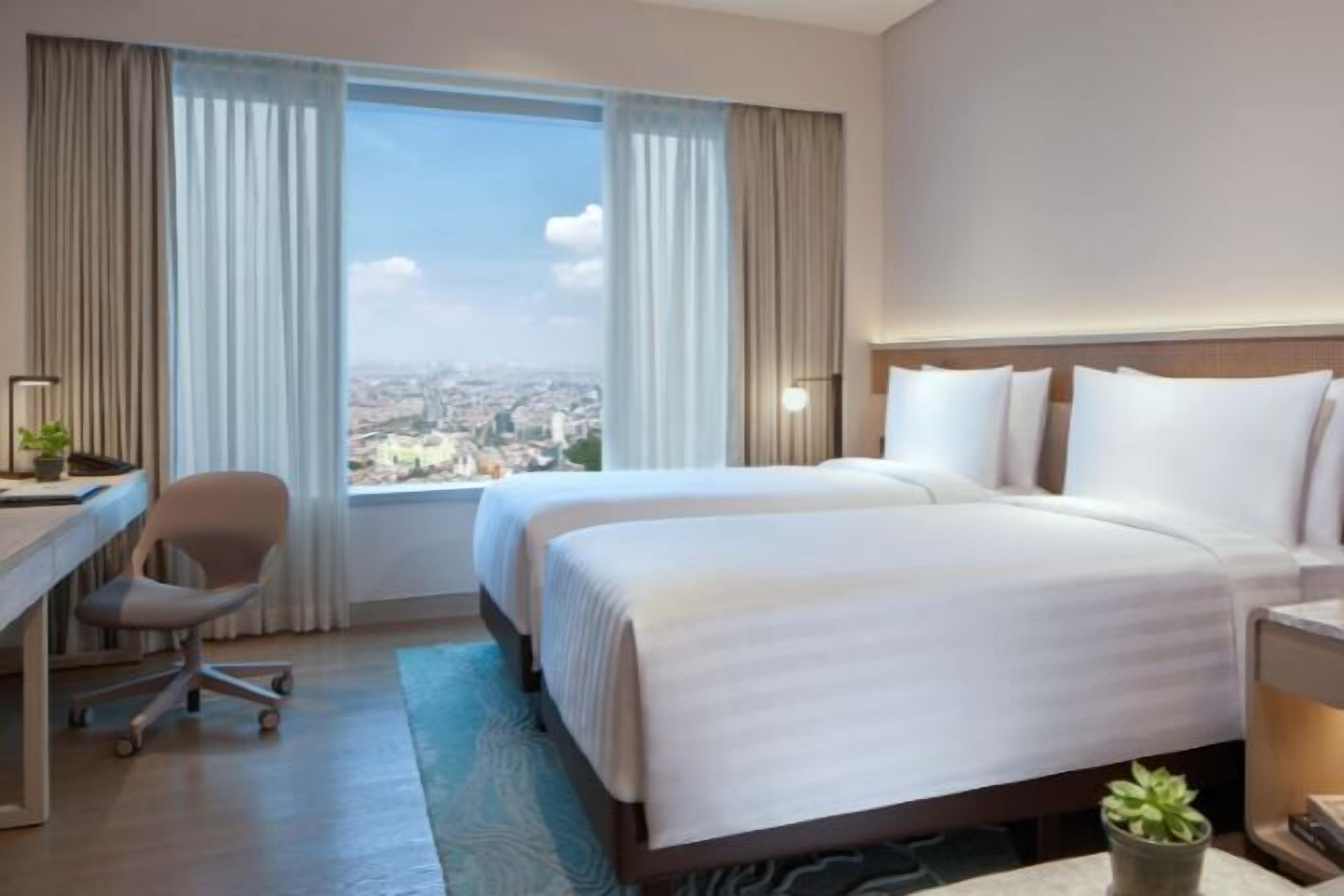 A hotel room with two beds, a desk, a chair, a small plant, and a view of the city.