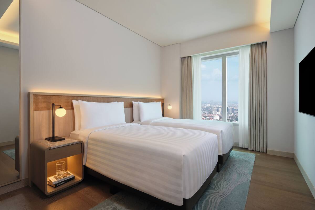 Habitación de hotel con dos camas, una mesita de noche con lámpara, un libro y vista a la ciudad a través de amplias ventanas.