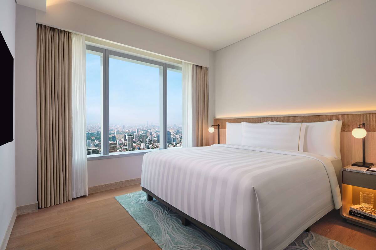 Habitación de hotel con una cama grande, una mesita de noche con lámpara y vistas de la ciudad a través de un ventanal, con un paisaje urbano visible desde el exterior.