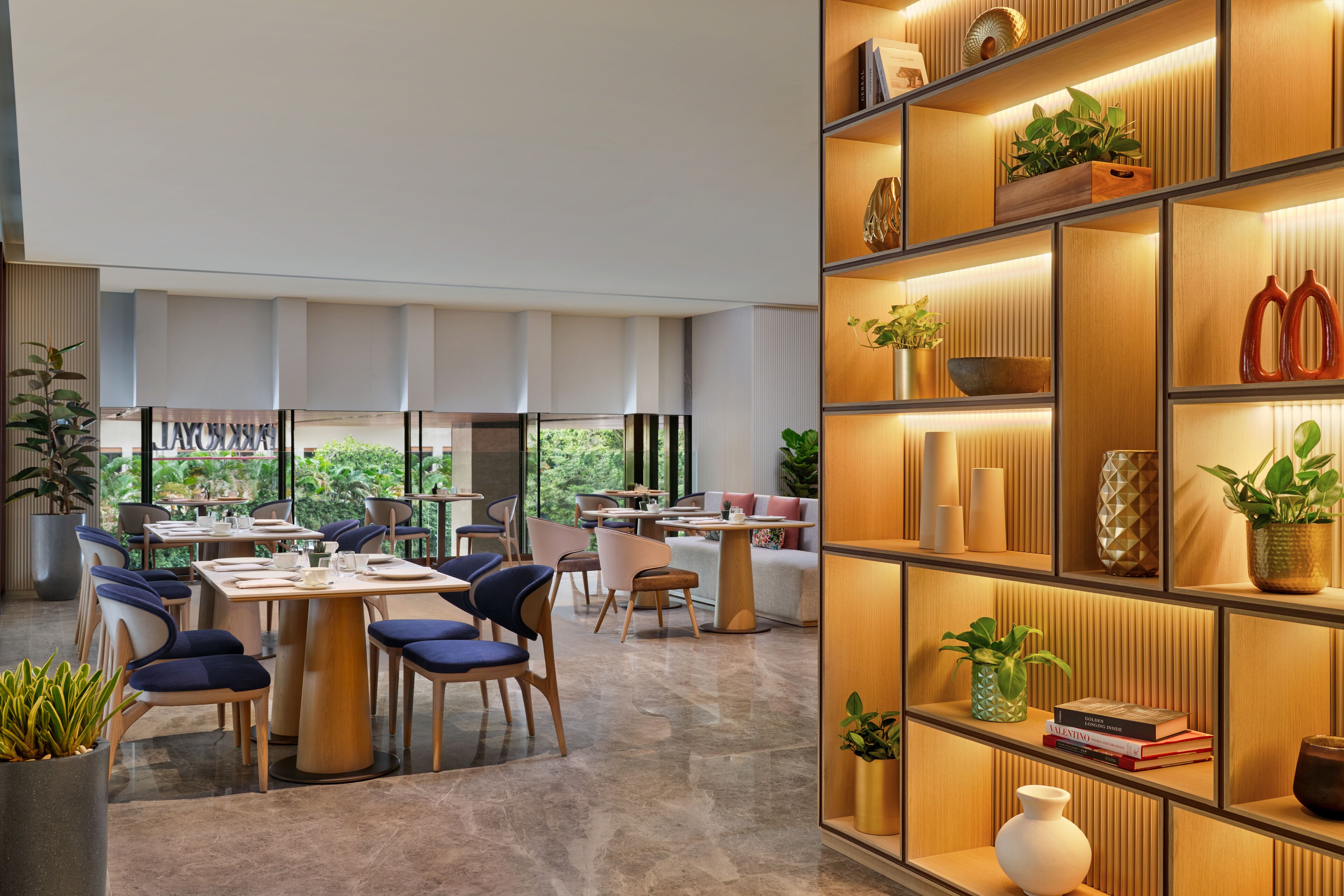 A modern dining area with tables and chairs, a bookshelf with decorative items, and a view of greenery outside.