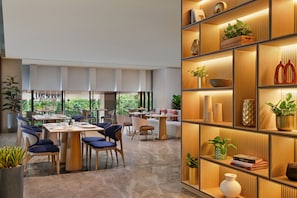 A modern dining area with tables and chairs, a bookshelf with decorative items, and a view of greenery outside.