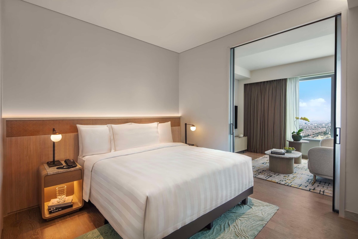 A modern hotel room with a large bed, bedside lamps, a nightstand, a small table, and a view of the city through a large window.