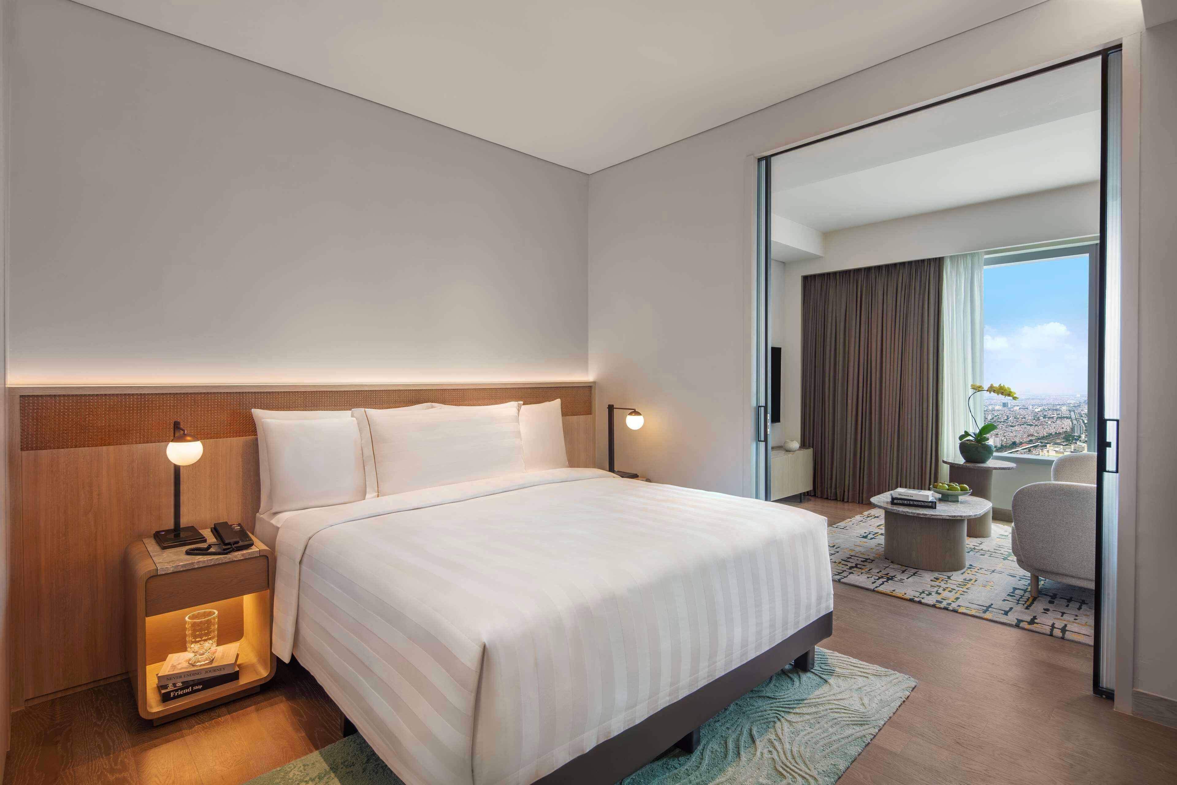 A modern hotel room with a large bed, bedside lamps, a nightstand, a small table, and a view of the city through a large window.