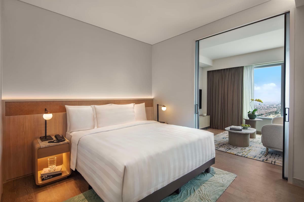 A modern hotel room with a large bed, bedside lamps, a nightstand, a small table, and a view of the city through a large window.