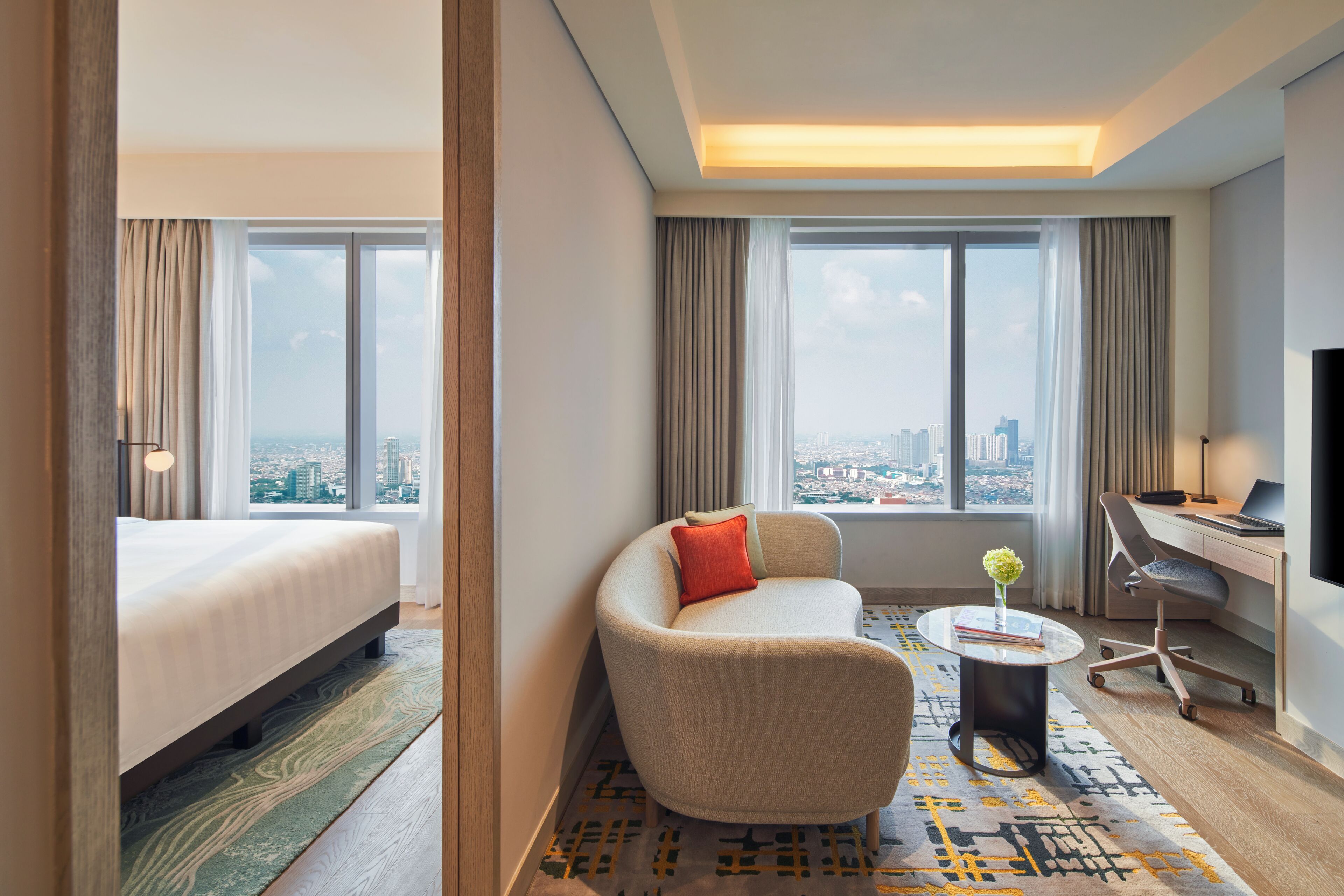Ein Hotelzimmer mit Bett, Sofa, Schreibtisch, Stuhl, Fernseher und großen Fenstern mit Blick auf die Stadt.