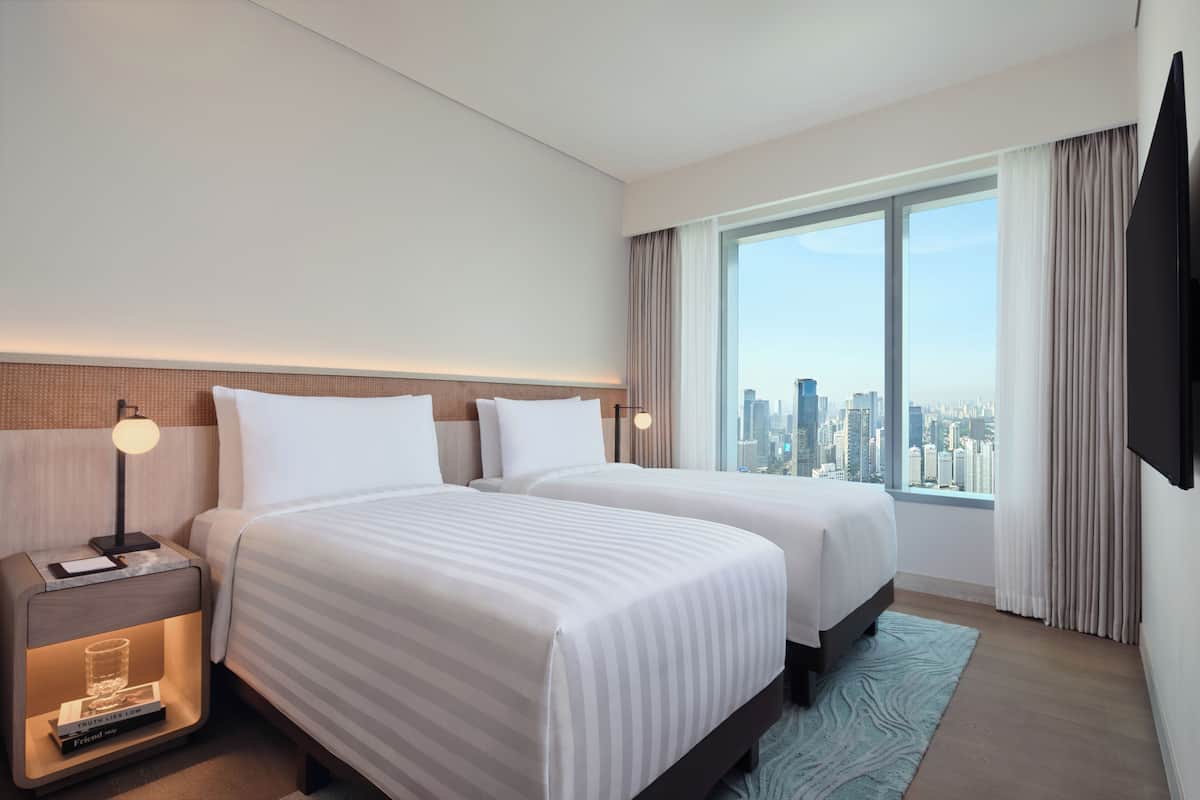 Habitación de hotel con dos camas, una mesita de noche, una lámpara, un ventanal con vistas a la ciudad y un televisor de pantalla plana montado en la pared.