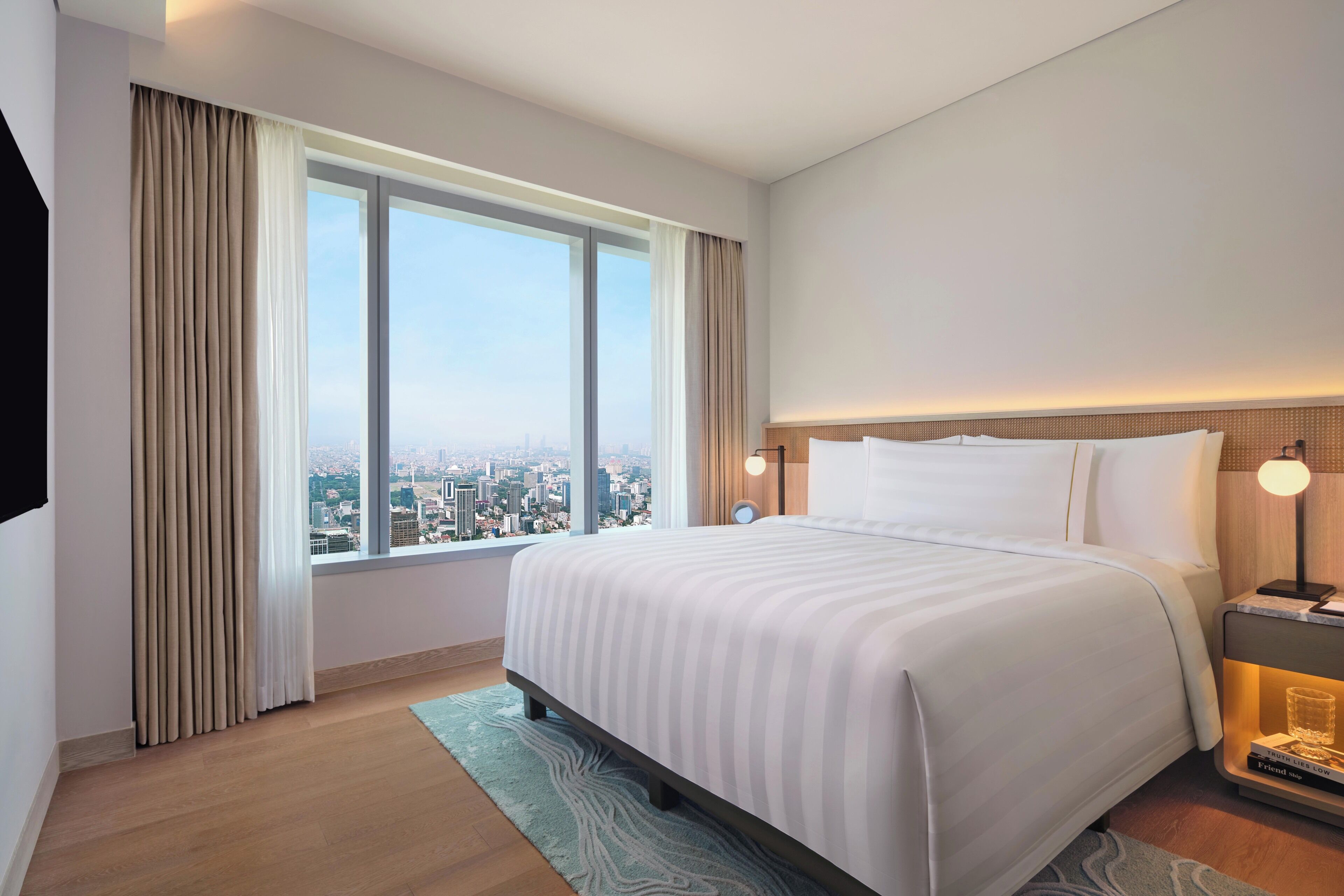 A hotel room with a large bed, a nightstand with a lamp, a view of the city through the window, and a cityscape visible outside.