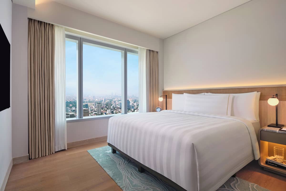 Un cuarto de hotel con una cama grande, una mesita de noche con lámpara, vista a la ciudad por la ventana y un paisaje urbano visible desde el exterior.