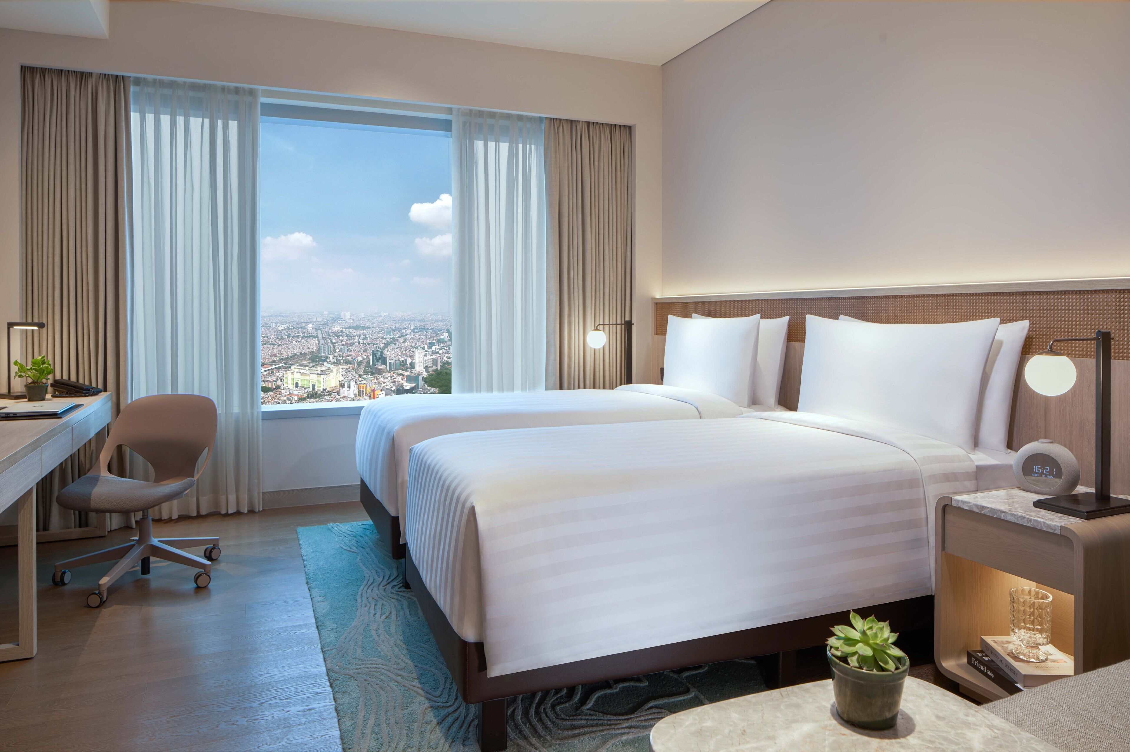 Ein Hotelzimmer mit zwei Betten, einem Schreibtisch, einem Stuhl, einem Nachttisch mit Lampe und Blick auf die Stadt.