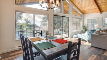 A dining area with a wooden table, chairs, and a chandelier. There are large windows and a view of the outdoors.