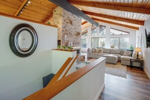 A spacious living area with a fireplace, a staircase with wooden railings, and large windows allowing natural light. - Tree-lined Gold Beach Retreat w/ Multiple Decks! (Gold Beach)