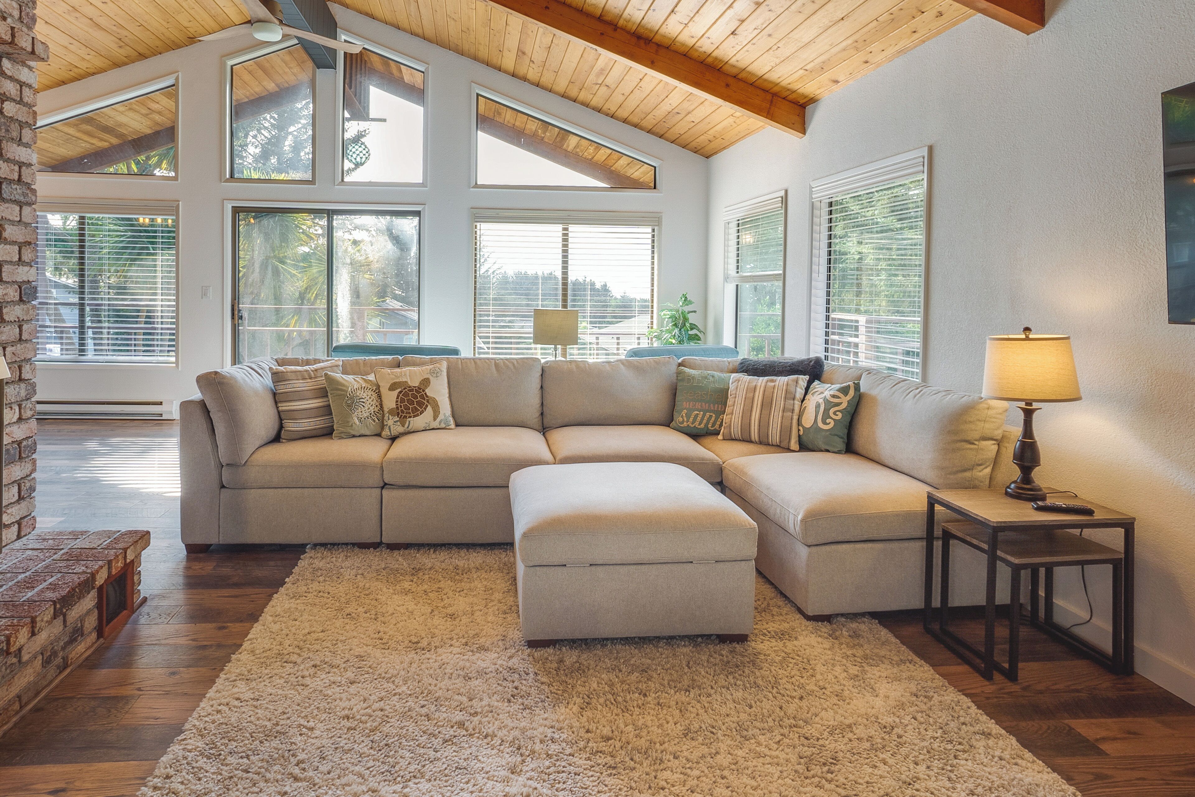 A sectional sofa with an ottoman, a small table with a lamp, and large windows with blinds.