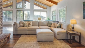 A sectional sofa with an ottoman, a small table with a lamp, and large windows with blinds.