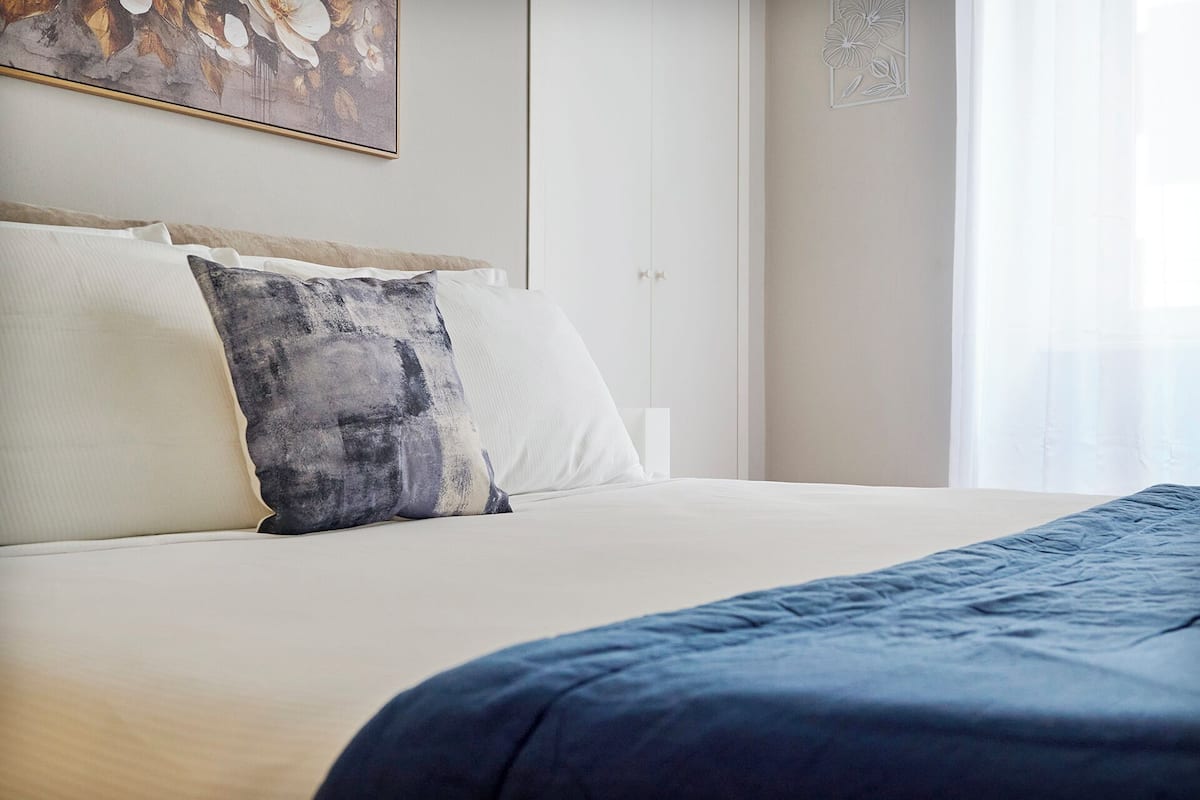 Een net opgemaakt bed met een blauwe deken, witte kussens en een grijs abstract kussen.