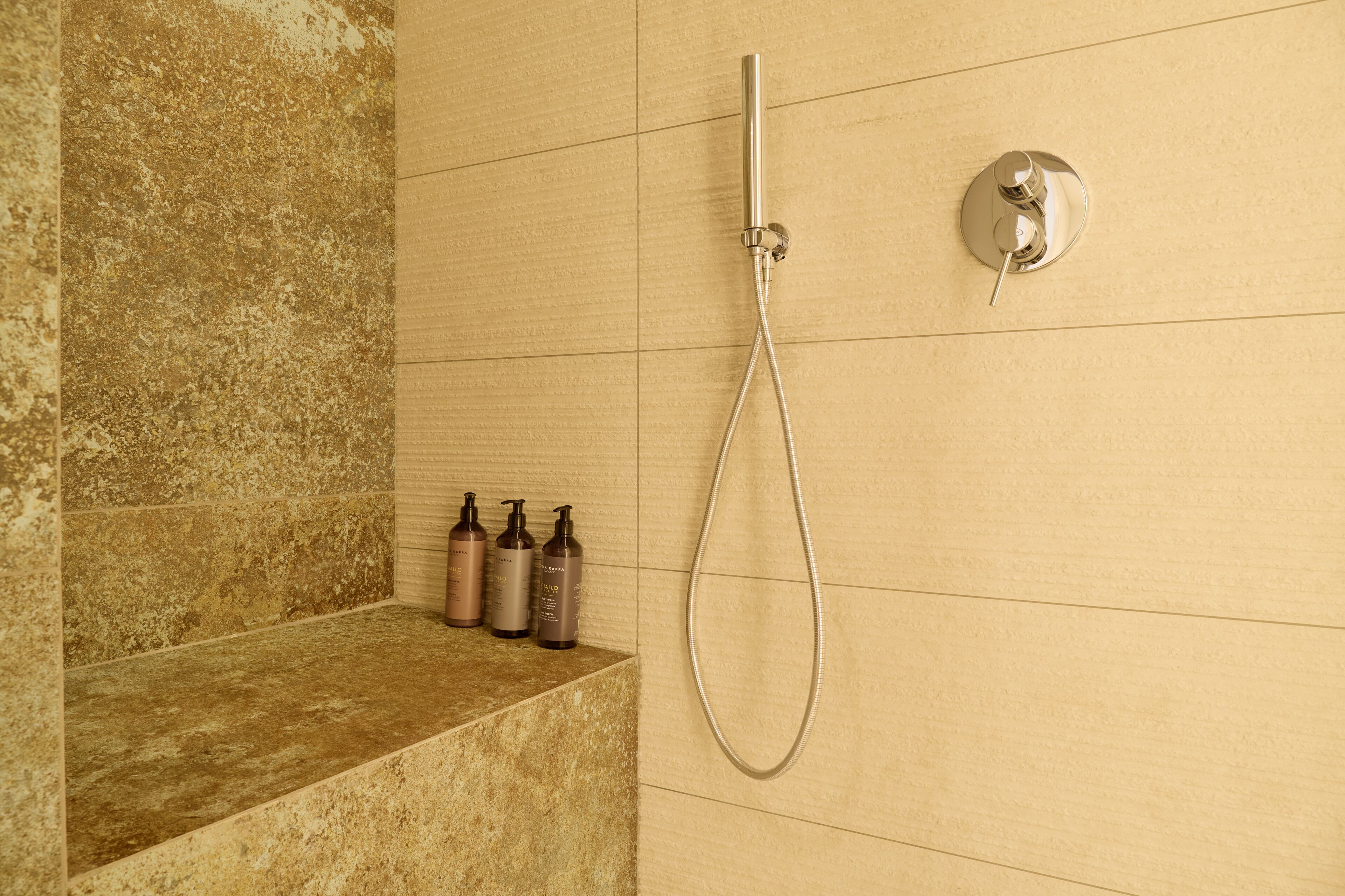 Un espace douche équipé d’un pommeau de douche à main et d’un panneau de commande fixé au mur. On y trouve trois bouteilles de shampoing et d’après-shampoing sur une étagère.