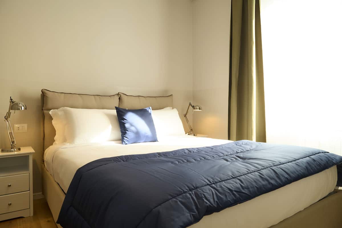 Een net opgemaakt bed met een blauwe deken en witte lakens, een nachtkastje met lampen en een raam met gordijnen.