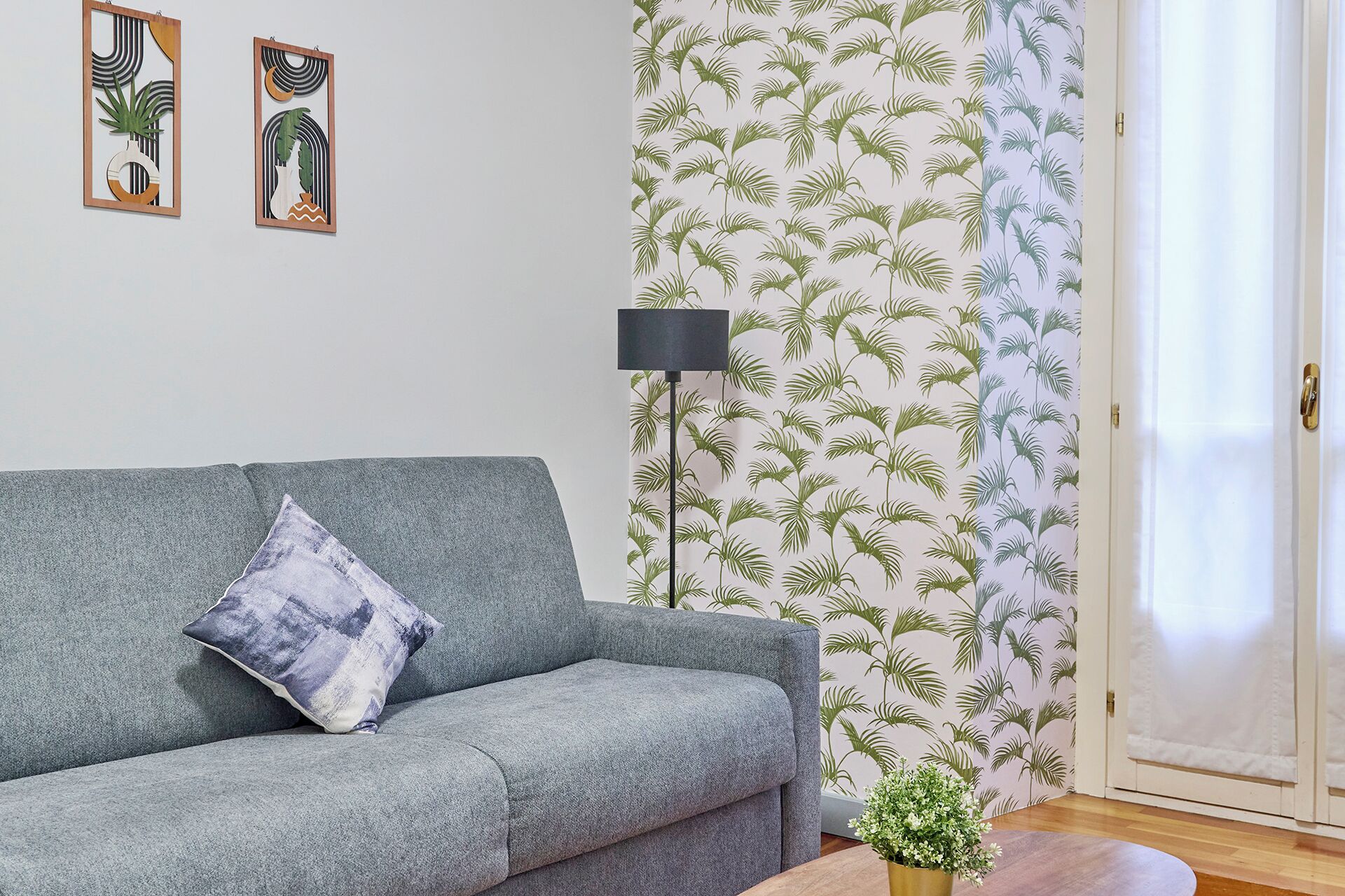 Uma sala de estar com um sofá cinza, uma mesinha com uma planta, um abajur e uma porta com cortina branca.
