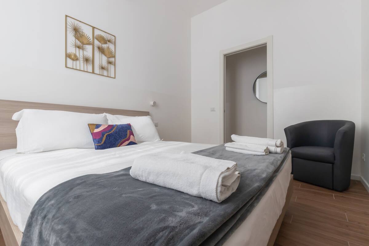Een net opgemaakt bed met witte en grijze lakens, een blauw en roze kussen en een grijze plaid. Een zwarte stoel staat rechts van het bed en een deur leidt naar een andere kamer.